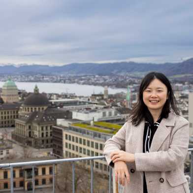 Yu Wang an einem Geländer lehnend, hinter ihr die Aussicht auf das ETH und Uni Gebäude und den Zürichsee. Der Himmel ist wolkenbehangen.