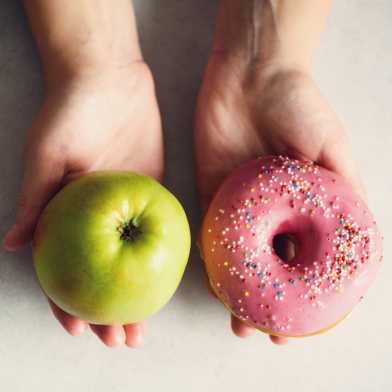 Eine Person präsentiert auf der einen Hand einen Apfel, auf der anderen einen Donut.