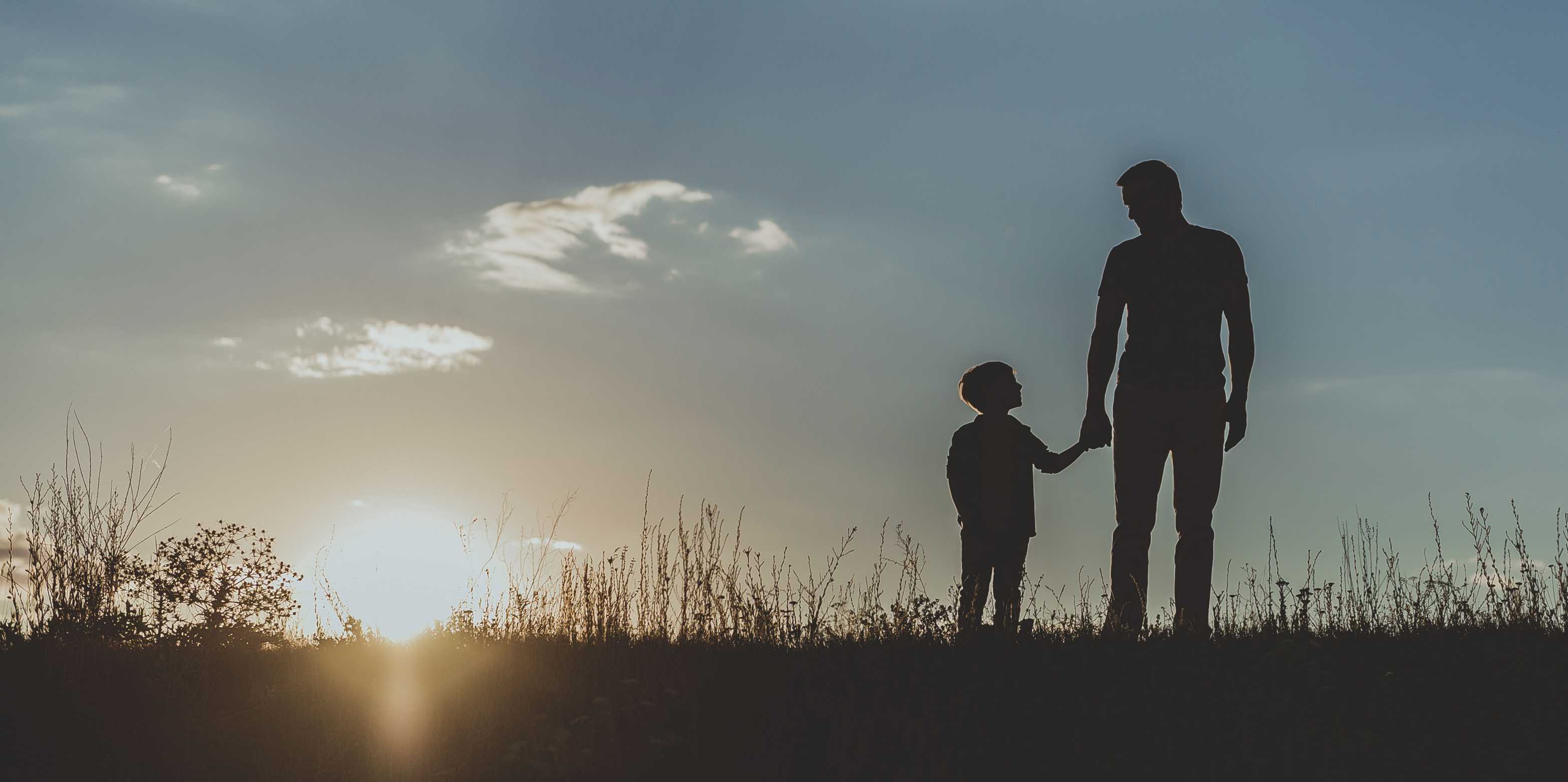 Sonnenauf- oder untergang im Hintergrund, im Vordergrund Vater und Sohn auf einer Wiese sich die Hand haltend