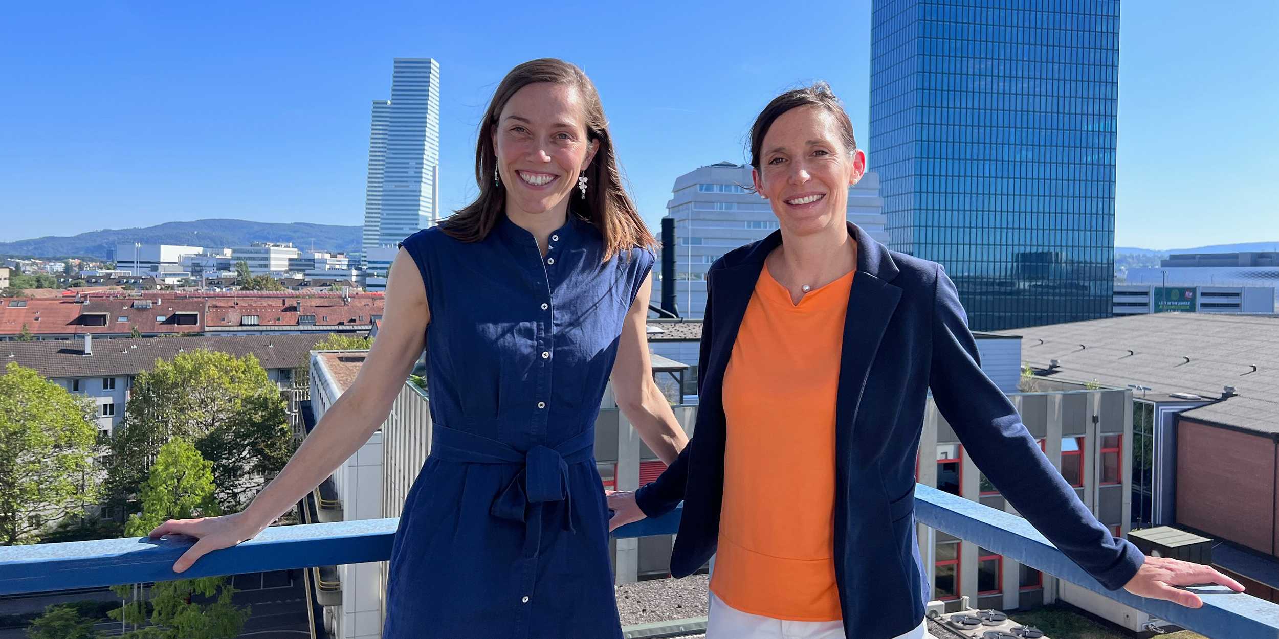 Bild von Barbare Treutlein und Tanja Stadler, die mit einem Lächeln im Gesicht an einem Geländer lehnen. Im Hintergrund sind Gebäude der Stadt Basel zu sehen.