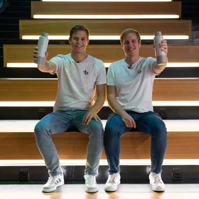 Christian Käser und Linus Lingg sitzen auf einer Treppe und halten ihre Bottleplus Flasche in die Höhe.