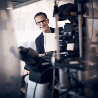 Dimos Poulikakos in seinem Labor schaut lächelnd hinter seinen Geräten hervor.