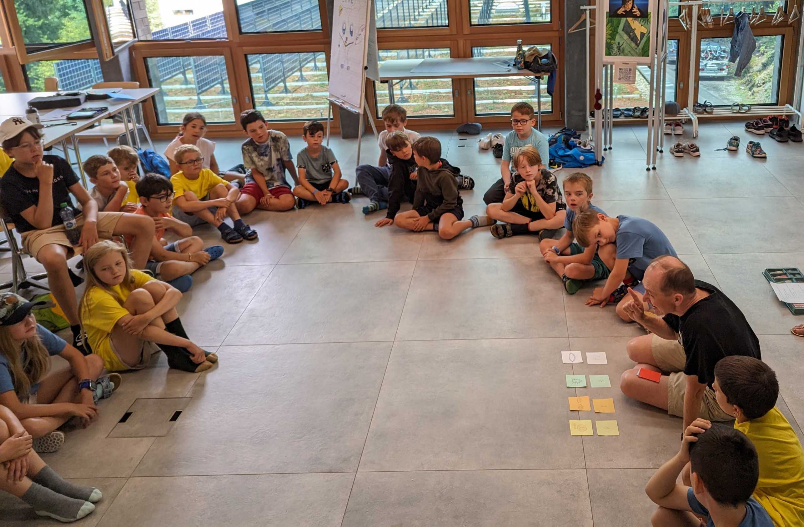 Viele Kinder sitzen in einem Kreis und hören den Erklärungen von Juraj Hromkovic zu.
