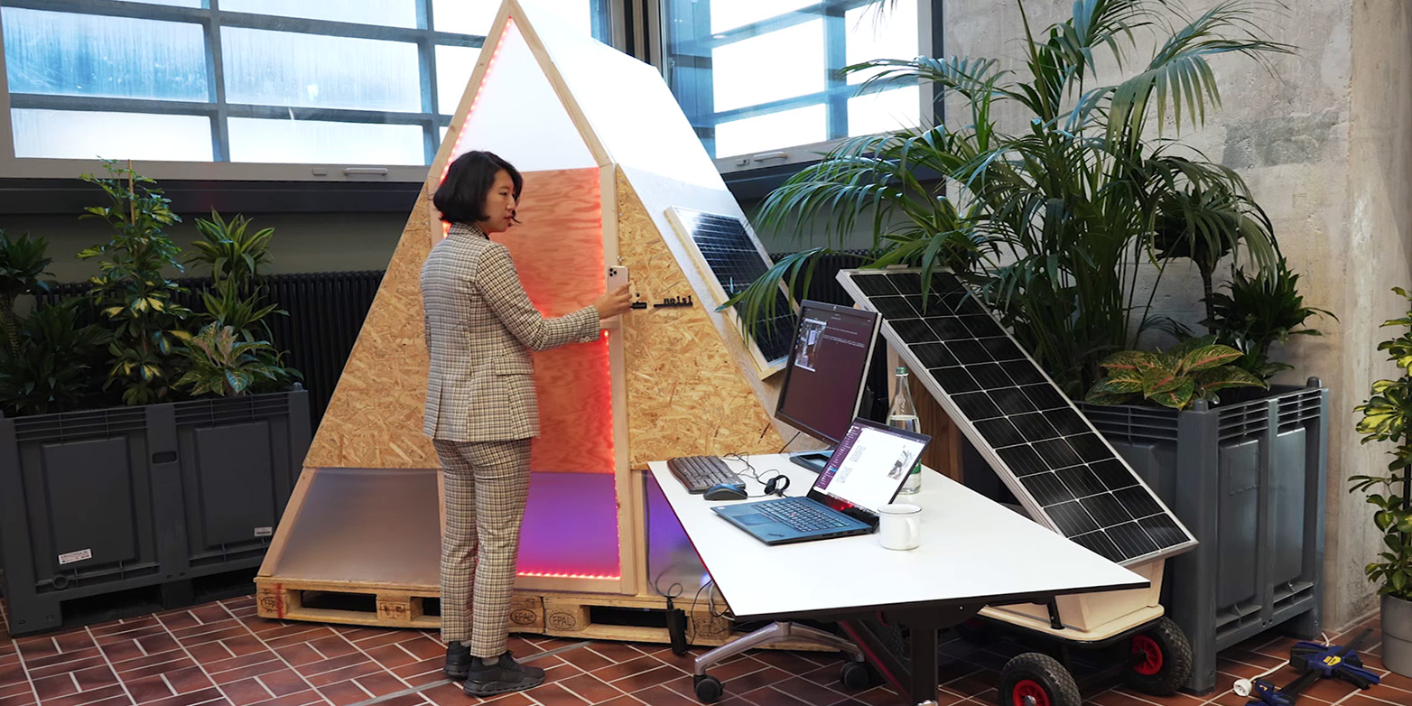 A woman stands in front of a triangular box with laptop and solar panel