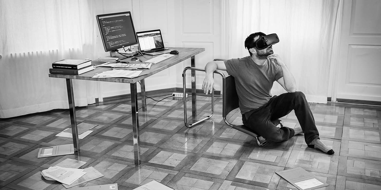 A man with VR glasses sits in a room on the floor, next to him is a desk with a computer