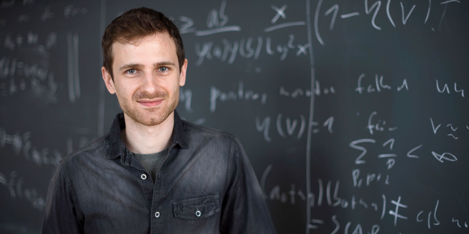 Oliver Janzer stands smiling in front of a written blackboard 