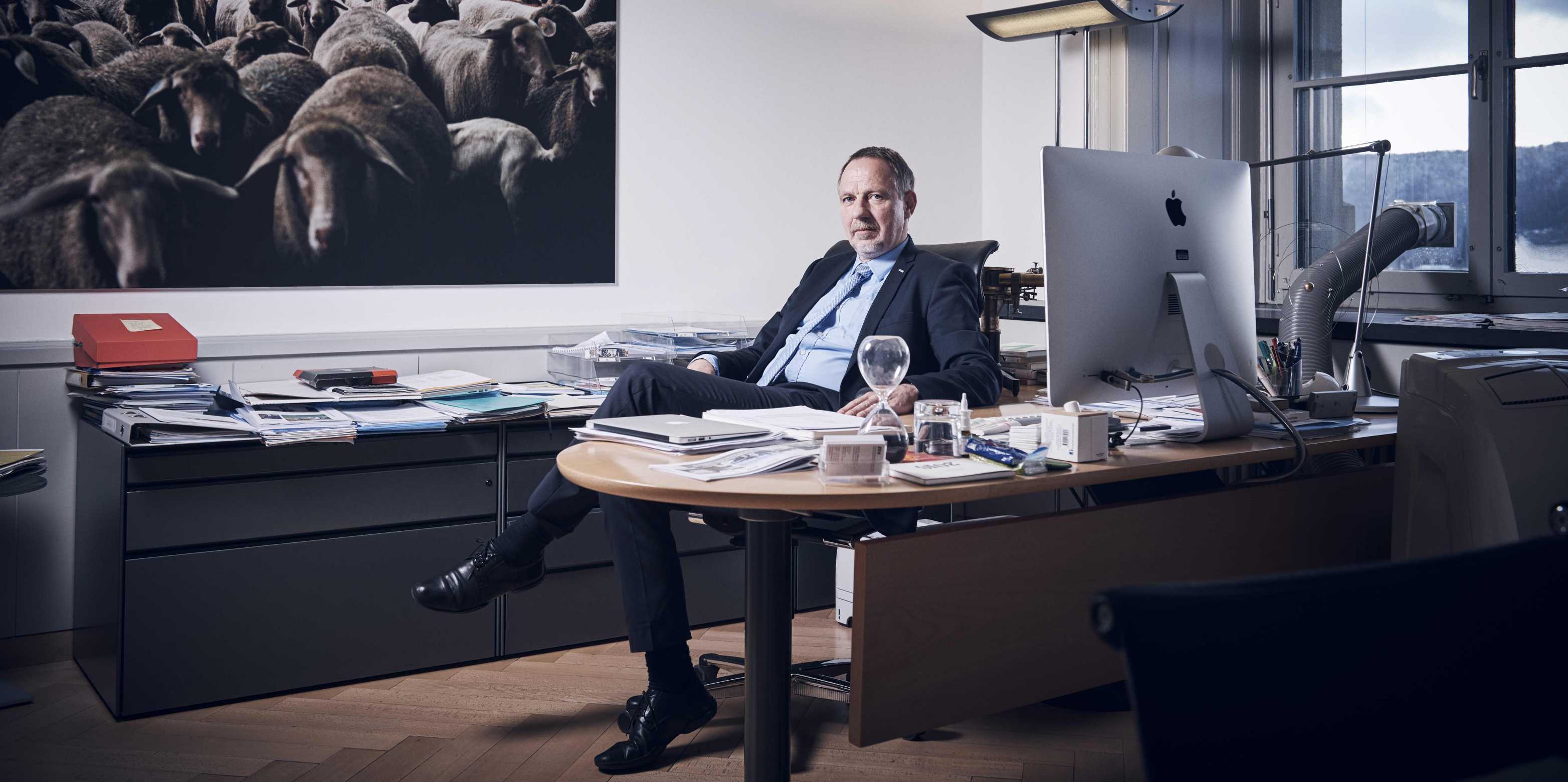 Detlef Günther is sitting in his office in a suit. In the back, there is a picture of a flock of sheep.