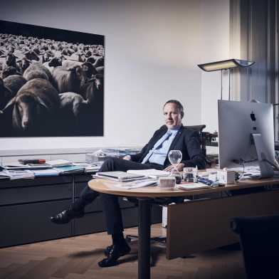 Detlef Günther is sitting in his office in a suit. In the back, there is a picture of a flock of sheep.