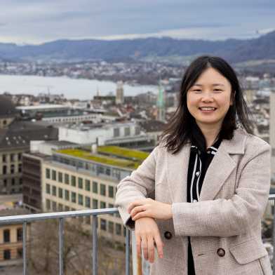 Yu Wang leaning against a railing, behind her the view of the ETH and Uni building and Lake Zurich. The sky is covered in clouds.