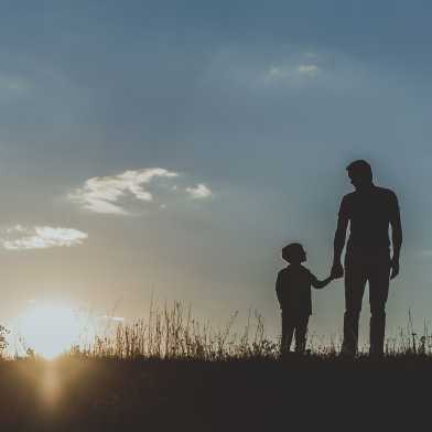 Sunrise or sunset in the background, in the foreground father and son holding hands in a meadow.