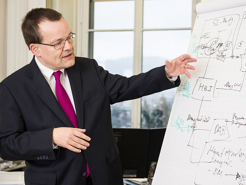 Enlarged view: Perich in front of a whiteboard, explaining which financial procedures are to be seen.