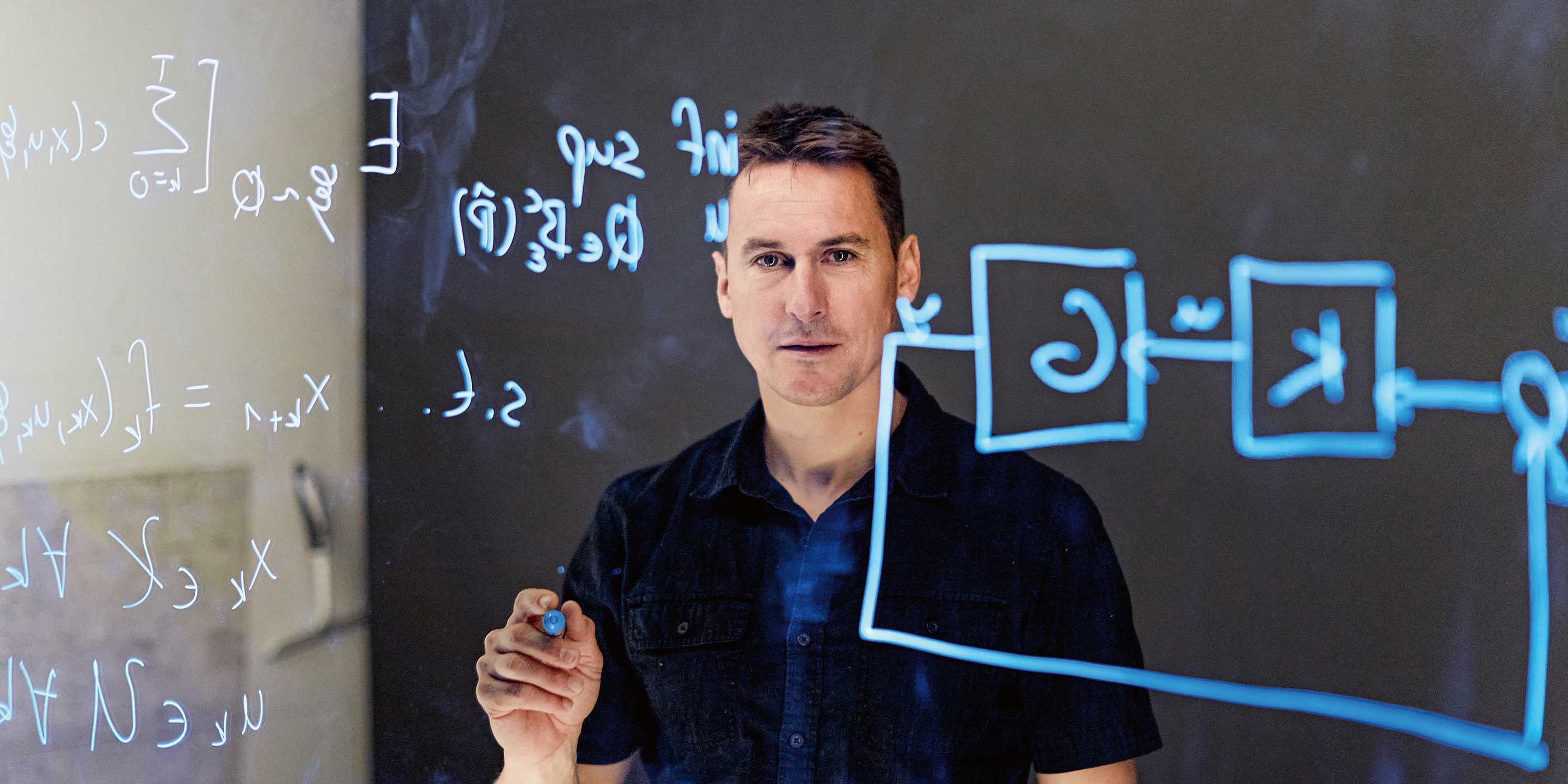 Florian Dörfler behind a glass panel with formulas in blue writing on it.