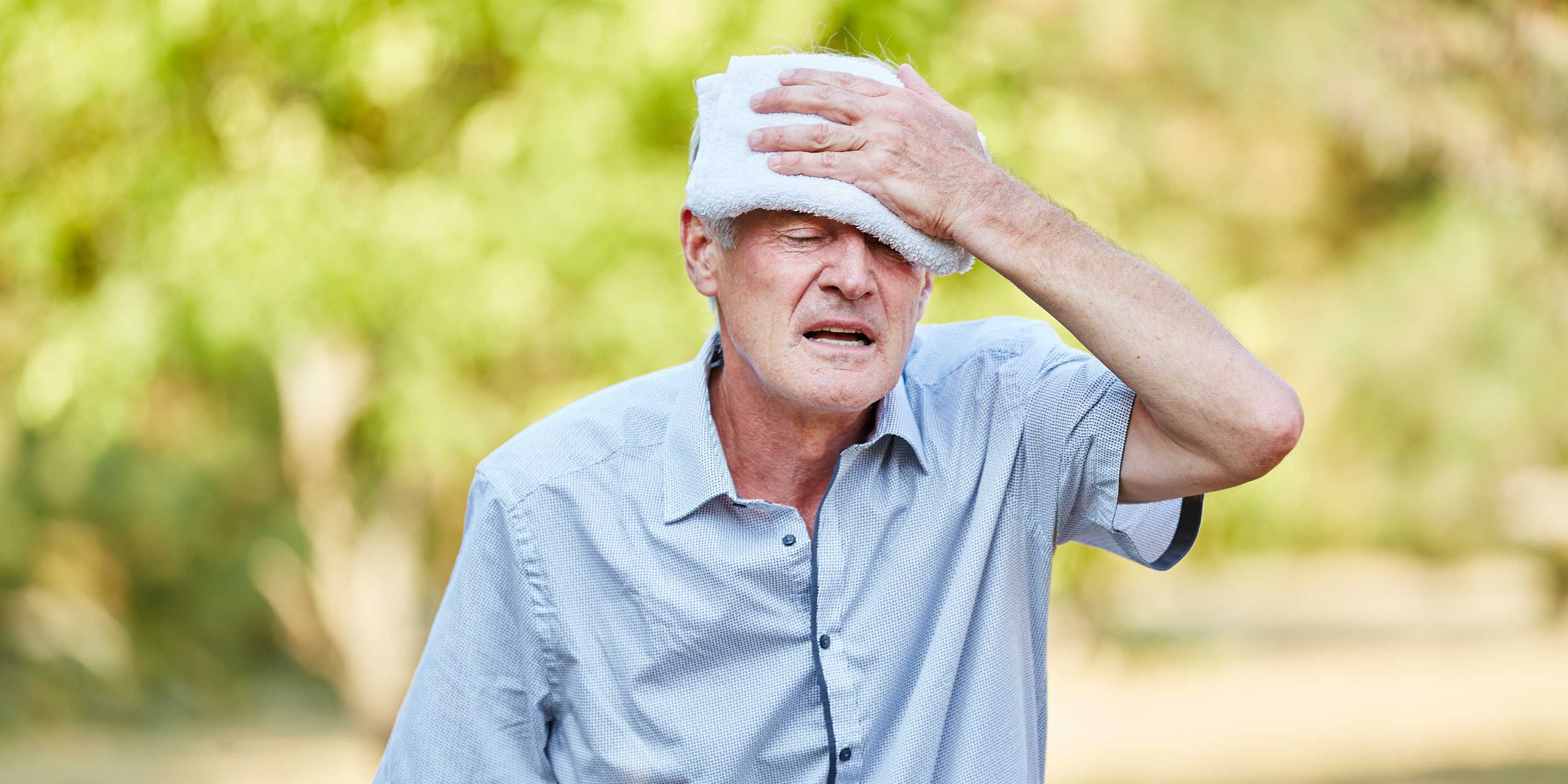 An old man holds a damp cloth to his forehead and contorts his face in suffering.