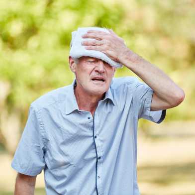 An old man holds a damp cloth to his forehead and contorts his face in suffering.