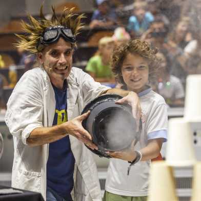 A scientist holds a device with a boy with which they aim at a paper cup tower.