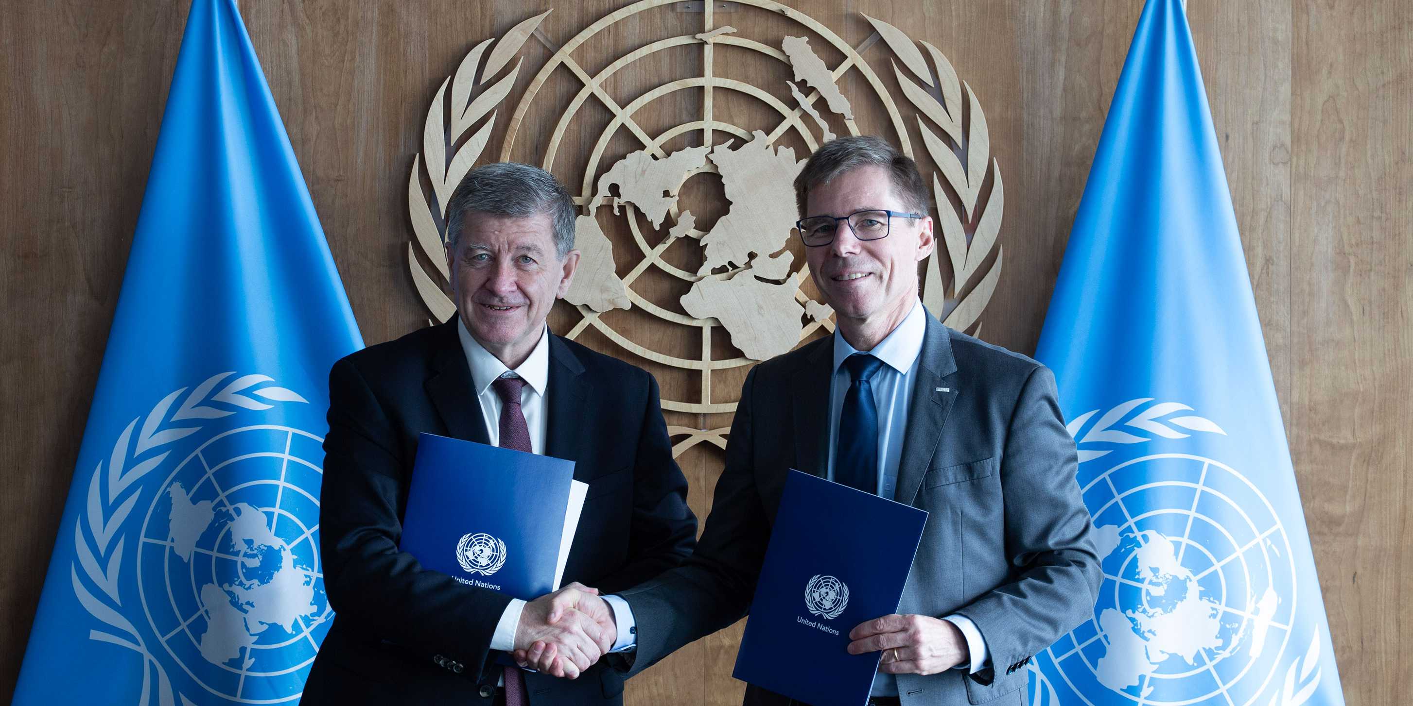 Guy Ryder, on the left, and Joël Mesot shake hands. They are each holding a blue folder with the UN emblem in the background.