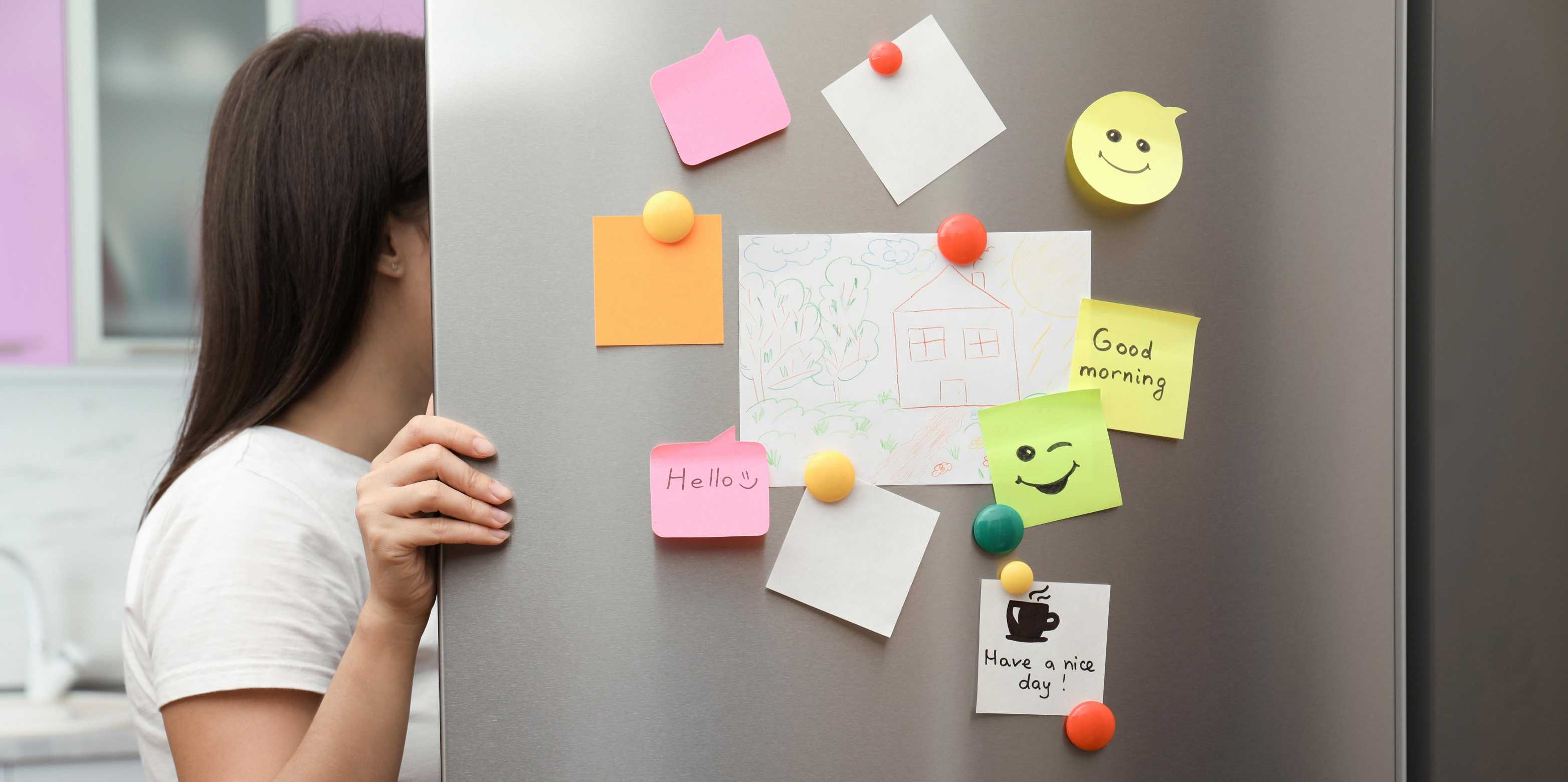 A woman opens the fridge and looks inside. The fridge door is covered in magnets and post-its.
