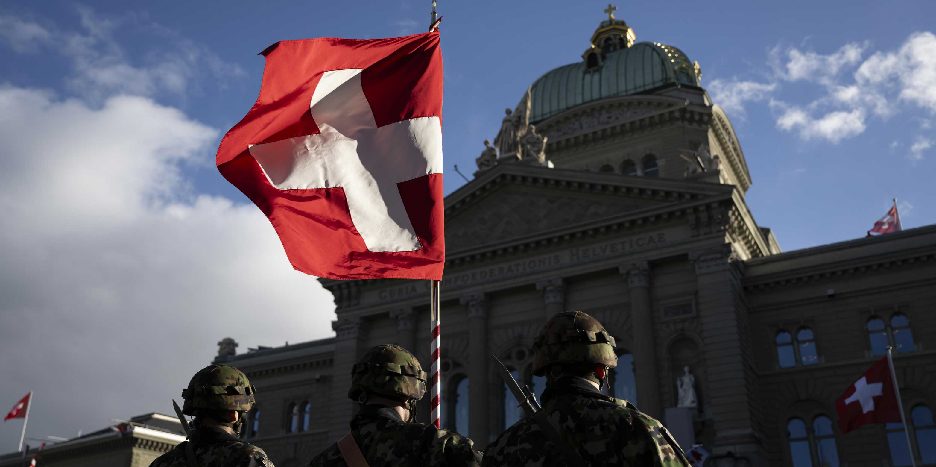 Three people in military uniform stand in front of the Federal Palace with a Swiss flag.